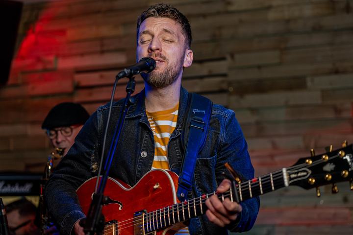 Tom Ryder in a denim jacket singing under red stage light during an evening party set