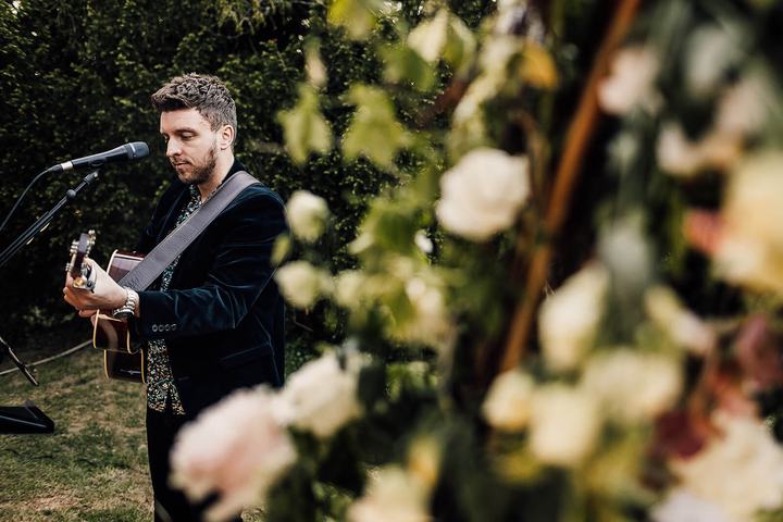 Tom Ryder in a dark suit playing beside a large white floral wedding arch