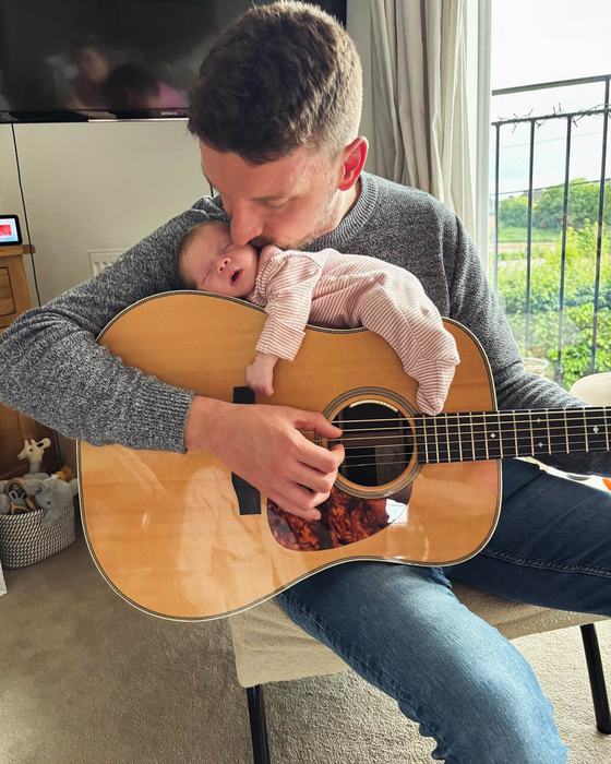 Tom Ryder at home holding his newborn baby against his acoustic guitar