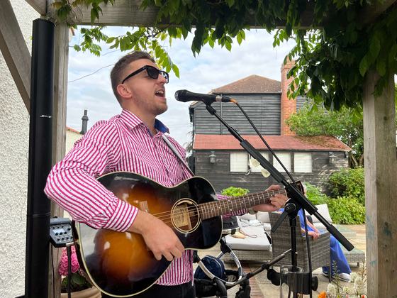Tom Ryder playing guitar outdoors under a pergola in a pink striped shirt