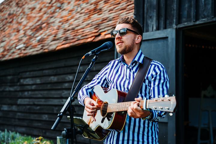 Tom Ryder outside a tiled barn wearing sunglasses, guitar in hand