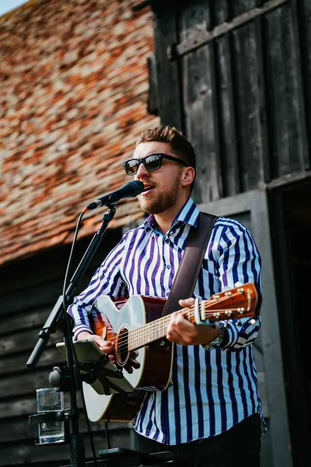 Tom Ryder singing and playing guitar outside a black timber barn at a wedding venue.