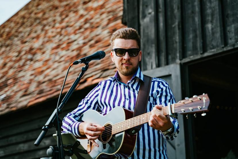 Tom Ryder singing and playing guitar outside a black timber barn, wide frame.