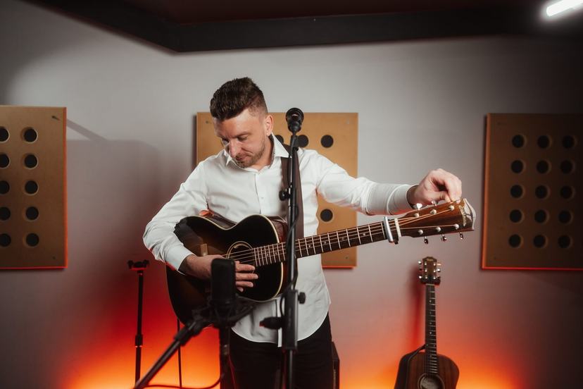 Tom Ryder tuning an acoustic guitar in a studio room, a microphone in front of him.